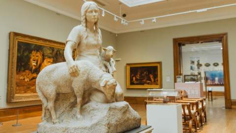 Marble sculpture of a figure with a sheep and a small animal on the figure's shoulder displayed in the spacious Ferens art gallery, Hull,  surrounded by classic framed paintings and wooden seating
