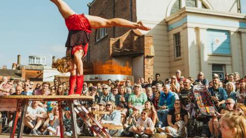 A woman in a red leotard is performing a hand stand on a raised platform with a crowd surrounding her.