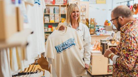 A customer browses a gift shop on Humber Street in Hull while holding up a Yorkshire‑themed T‑shirt, surrounded by shelves of local souvenirs and merchandise.