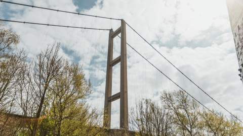 View of the Humber Bridge in Hull framed by trees, showing the tall suspension tower and cables against a bright, cloudy sky.