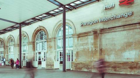 Exterior view of Hull Paragon Interchange showing its historic arched windows, sandstone façade and station signage, with blurred commuters walking through the covered concourse.