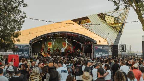 The crowd at Humber Street Sesh at sunset. The crowd are wearing summer clothes and are watching a band at the Hull Trains stage which is branded in the blue Hull Trains logo with hearts. Behind the stage, The Deep's iconic sloping arcitecture shines in the sunset.