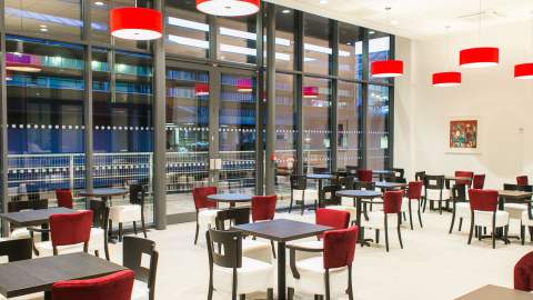 Bright and modern café at Ferens Art Gallery in Hull featuring red pendant lighting, large floor‑to‑ceiling windows, and neatly arranged tables and chairs. The spacious, contemporary setting highlights the gallery’s welcoming visitor facilities