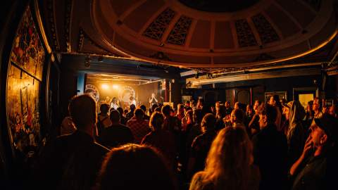A large crowd of people in the Polar Bear stands inside a dimly lit music venue, facing a brightly lit stage where a band is performing, with warm lighting casting an orange glow over the audience and ornate details visible on the ceiling above.
