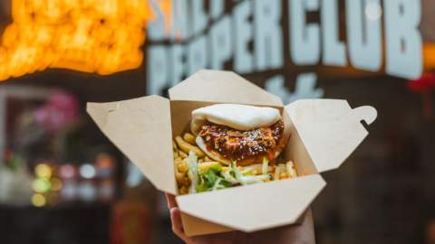 Takeaway box filled with seasoned fries, salad and a bao bun in front of the neon sign shop front of Salt and Pepper Club in Paragon Arcade