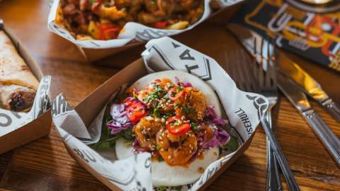 Close‑up of street‑food style dishes in Hull, including a bao bun with crispy prawns, slaw and chilli, served in cardboard trays on a wooden table