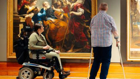 A powered wheelchair user and another visitor with a mobility aid admire the  large classical painting 'Tobias Healing the Blind Tobit' (c.1650) by Valerio Castello in the spacious, accessible Ferens art gallery Hull .