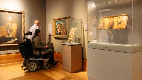 A visitor using a powered wheelchair explores the Renaissance gallery at Ferens Art Gallery in Hull, viewing illuminated religious panels and framed paintings displayed in glass cases. The scene highlights the gallery’s accessible layout and its collection of historic Renaissance artwork