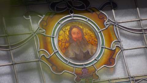 Decorative stained‑glass window featuring ornate metalwork and a circular central panel with an illustrated historic figure, surrounded by detailed patterns and warm coloured glass.