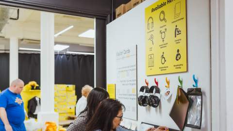 Visitors at an accessibility support station in a Hull venue, viewing a wall display offering sensory packs, ear defenders and other accessible resources, with staff and equipment visible in the background.