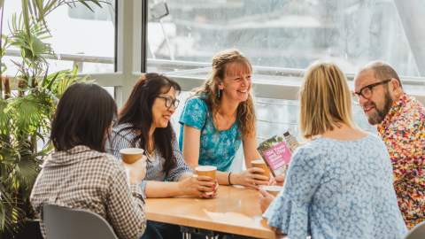 A small group of people sitting together in a bright café space at The Deep in Hull, enjoying hot drinks and discussing visitor guides near large windows and indoor plants