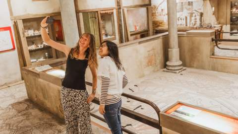 Two visitors explore a historic Roman‑inspired gallery in Hull, taking a photo together beside detailed archaeological displays and reconstructed ancient buildings. The scene highlights Hull’s accessible cultural attractions and immersive heritage exhibits.