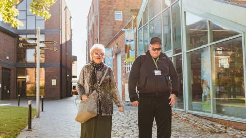 Two people walking along a cobbled street in Hull’s Museums Quarter, surrounded by brick buildings, greenery and large glass-fronted museum entrances on a sunny day.