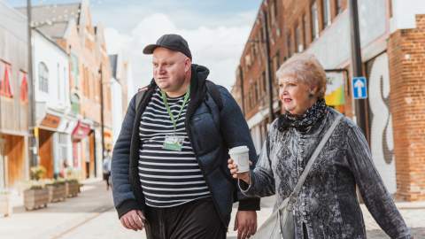 Two people walking through Hull's fruit market , holding a takeaway coffee and passing brick buildings, independent shops cafes and restaurants on a bright day