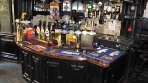 Traditional pub bar interior featuring polished wood counters, multiple beer pumps, glassware displays and an array of spirits and drinks behind the bar, showcasing a classic British pub atmosphere.
