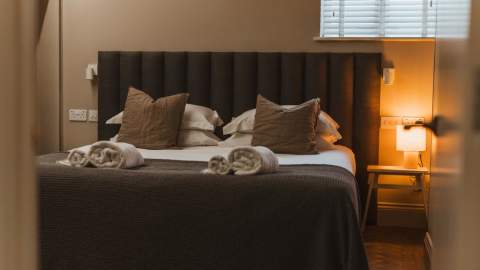 Hideout hotel bedroom. Cosy and inviting. The bedside lamp is on to the right of the bed. The bed looks soft and has a chocolate headboard, white sheets and brown accent cushions. There are fresh towels rolled up on the bed.