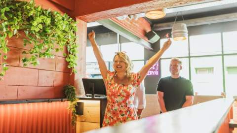Group playing darts in the orange panelling lanes of the Boardroom. A woman in a floral dress is celbrating.
