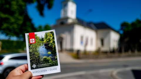 En hånd holder i en brosjyre for Borgleden foran en kirke