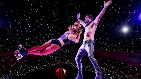Two people performing an acrobatic stunt. One person is wearing just trousers and no top whilst someone else is hanging around their neck as they spin around.