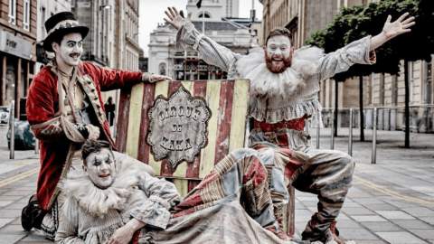 Three people dressed as clowns in grey and red clothing in front of a yellow and red sign that says Cirque du Kaka.