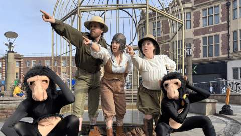 Five performers in a public square act out a scene in front of a large metal cage structure. Three in the centre wear vintage explorer-style clothing and point urgently into the distance with surprised expressions, while two others dressed in black bodysuits with ape masks crouch in the foreground, mimicking confused gestures.
