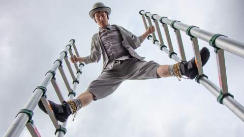 A performer dressed in grey with a grey hat on. They are standing between to ladders looking down at the camera.