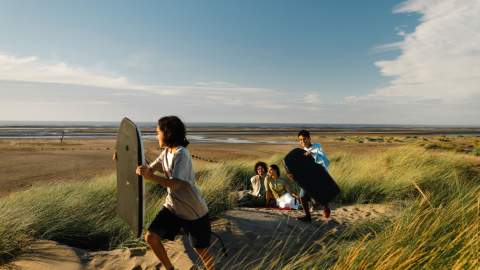 A family playing on sand dunes on a beach with small surf boards. There is a blue sky in the background.