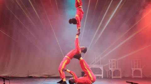 Three acrobats wearing red body suits. One is lying on the floor holding up another person with their legs. A third person is doing a hand stand on the second person's hands in the air.