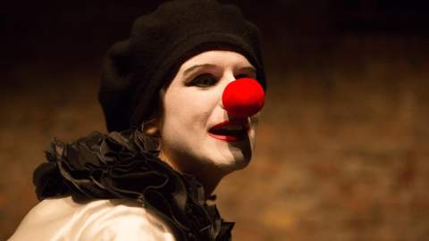 A person dressed as a clown, wearing white face makeup, a black beret, and a ruffled black collar, with a bright red clown nose. They are turned slightly to the side against a warm-toned background, with dramatic lighting highlighting their face.