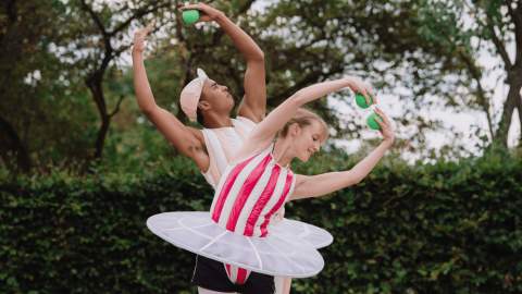 Two people in ballet outfits wearing tutus. They are posing with their arms in the air over their heads holding green balls.