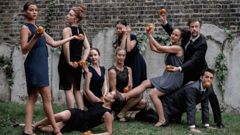A group of eight adults pose theatrically in a garden against a brick wall, all dressed in dark formal or semi-formal outfits. Each person is holding oranges in stylised, almost sculptural poses, some standing, some seated, and one lying on the grass. One man crouches on all fours with an orange in his mouth while another person balances across his back. The group’s expressions are serious and composed, creating a surreal, staged tableau.