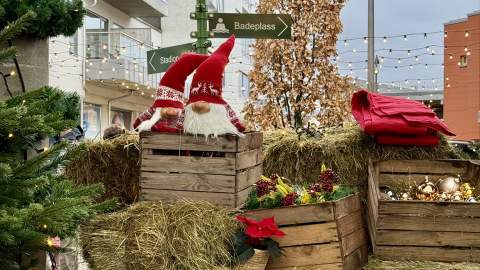 En juledekorert scene i sentrum av Lyngdal, med halmballer, trekasser fylt med julepynt, to nisser, røde julestjerner og et grønt veiviserskilt som peker til lokale attraksjoner. Små lyslenker og et tre i bakgrunnen gir førjulsstemning.