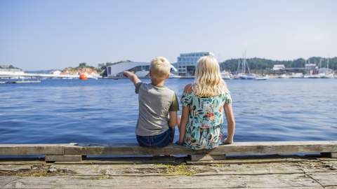 Two children sitting on a dock, pointing towards a building