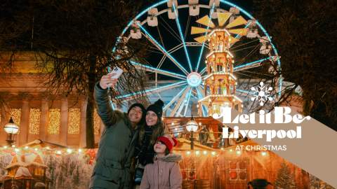 Two people taking a selfie photo with a child in front of them. They are standing in front of a Ferris wheel and a Christmas market.