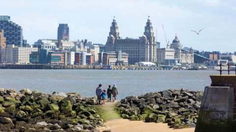 People stand on the shoreline surrounded by rocks looking over to the Liverpool skyline