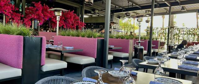 Colorful pink dining booths and wood tables are set for diners at the expansive, shaded patio at The Mexicano. The courtyard is filled with ample flowers and plants, creating an upscale, nature-filled dining room.