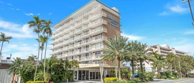 An eight-story hotel with a portico in front. Several palm trees and greenery line the drive