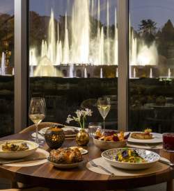 A table set for two with two champagne glasses and plates full with food. A window in the background shows Longwood Garden's fountains on display.