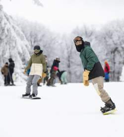 A group of people in snow gear snowboarding at Laurel Mountain Ski Resort. Snow covered trees can be seen in the background