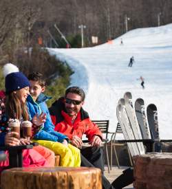 A family of four sitting at a firepit with the skis leaning up against the pit. People in the background can be seen skiing down a snowy hill.