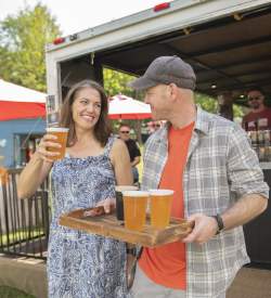 A man carrying a tray of beers and a woman walking with him with a beer in her hand at a festival