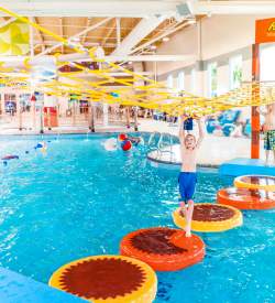 Young boy in a swim suit crossing floating pads using a net that hangs above him at Hershey Lodge's indoor waterpark.