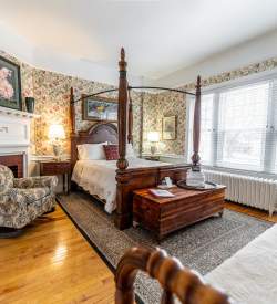 Interior of a bedroom with floral wallpaper in an historic inn with a full bed with a wooden headboard, a sitting chair, and a fireplace.