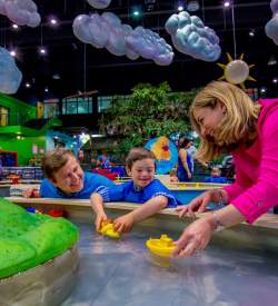 A mom, dad, and child playing with small toy boats in a stream display at a museum