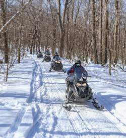 Four people snowmobiling in winter gear on an outdoor, snowy trail