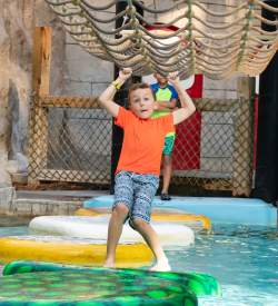 Little boy in a bathing suit and swim shirt using a roped net above him to cross floating pads in a pool