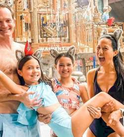 Family of four in bathing suits at an indoor waterpark posing for a photo. The one girl is being lifted up by her family
