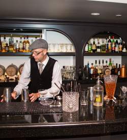 Man behind a bar with a cap on making a drink in a speakeasy