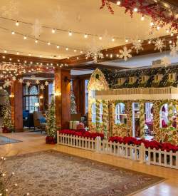 Interior of a hotel lobby adorned with holiday lights, pine trees, and a life-size house