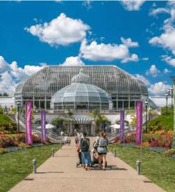 Two women each pushing strollers as they walk down a path framed with flower beds to the entrance of Phipps Conservatory